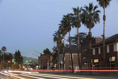Evening street scene in a small town with long-exposure light trails from passing cars, with palm trees lining the sidewalk.